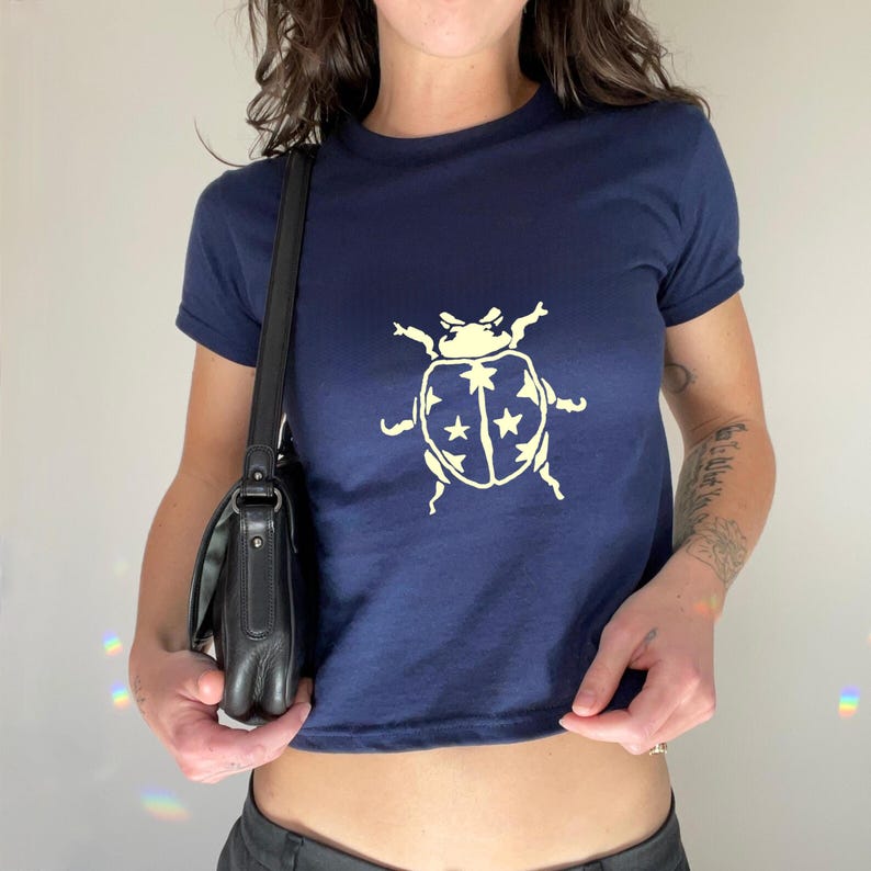 Op de afbeelding: Marineblauwe t-shirt met een cr&egrave;mekleurige lieveheersbeestje-afbeelding met sterren. De top met korte mouwen heeft een ronde hals. Het shirt is gemaakt van zacht materiaal.