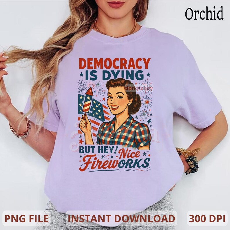 May include: A light purple t-shirt with a graphic featuring a vintage illustration of a woman holding a firework and American flag. The text reads "DEMOCRACY IS DYING BUT HEY! Nice Fireworks."