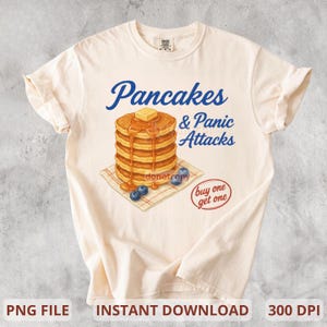 Può includere: T-shirt color crema con una pila di pancake con burro e sciroppo, mirtilli e la scritta "Pancakes & Panic Attacks". È presente anche una piccola grafica "buy one get one".