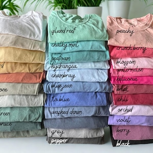 Pode incluir: Pilhas de camisetas de v&aacute;rias cores, incluindo branco, marfim, manteiga e mostarda. Outras cores incluem recife da ilha, menta giz, p&ecirc;ssego e cranberry. Cada pilha tem uma etiqueta escrita &agrave; m&atilde;o indicando o nome da cor.