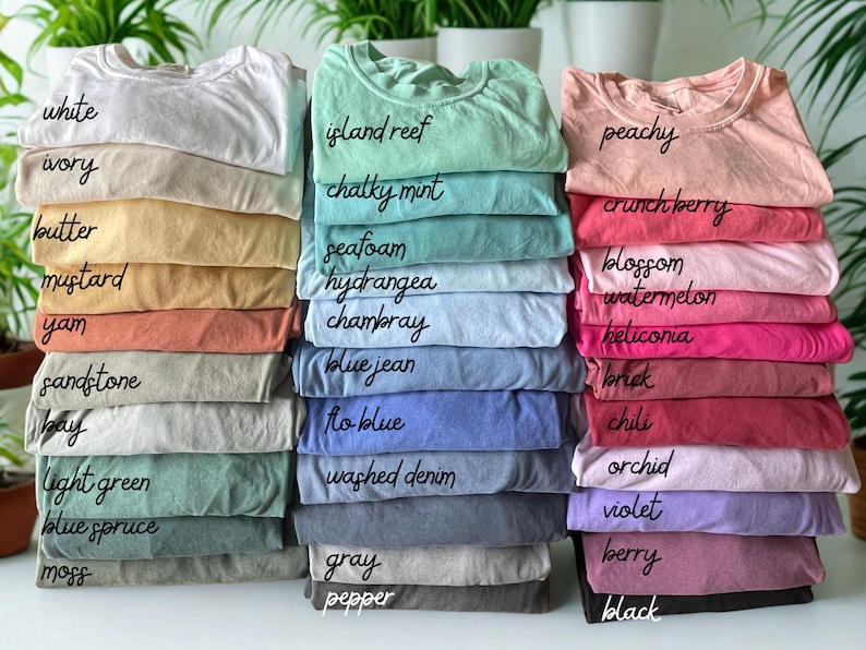 Pode incluir: Pilhas de camisetas de v&aacute;rias cores, incluindo branco, marfim, manteiga e mostarda. Outras cores incluem recife da ilha, menta giz, p&ecirc;ssego e cranberry. Cada pilha tem uma etiqueta escrita &agrave; m&atilde;o indicando o nome da cor.