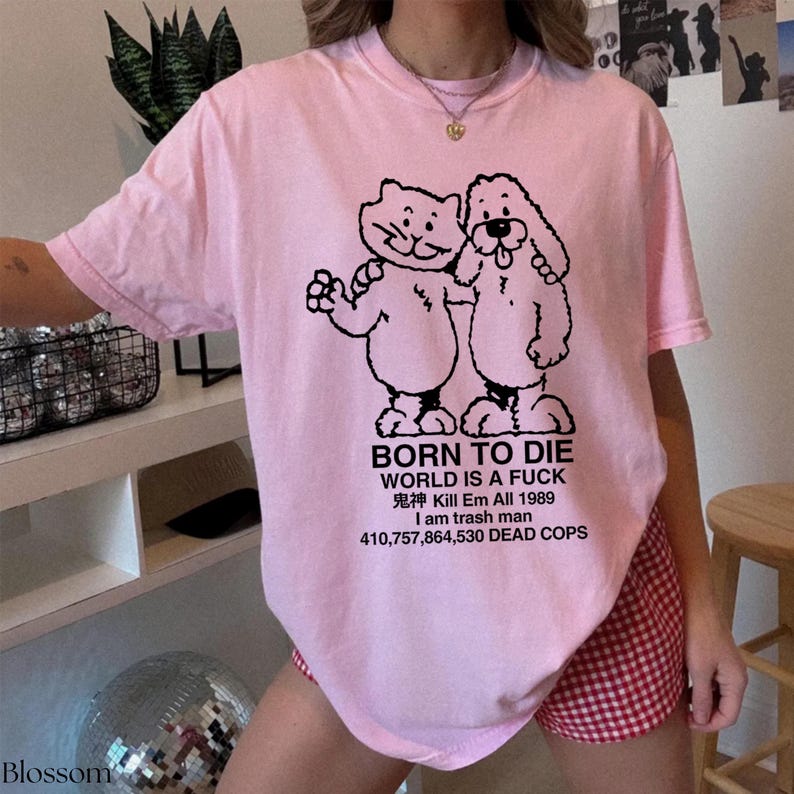 Pode incluir: Camiseta rosa claro com um desenho preto de um gato e um cachorro. Abaixo, o texto em preto diz: "BORN TO DIE WORLD IS A FUCK 鬼神 Kill Em All 1989 I am trash man 410,757,864,530 DEAD COPS". Roupa casual.