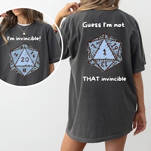 Puede incluir: Camisetas gris oscuro con un gráfico de dados D20 azul claro y marrón. Una camiseta dice "I'm invincible!" y la otra "Guess I'm not THAT invincible".