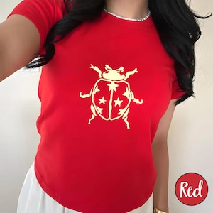 Op de afbeelding: Een felrood T-shirt met een cr&egrave;mekleurige lieveheersbeestjesafbeelding. Het lieveheersbeestjesontwerp heeft sterren op de rug. Het woord "Red" staat in een rode cirkel rechtsonder. Het shirt heeft korte mouwen.