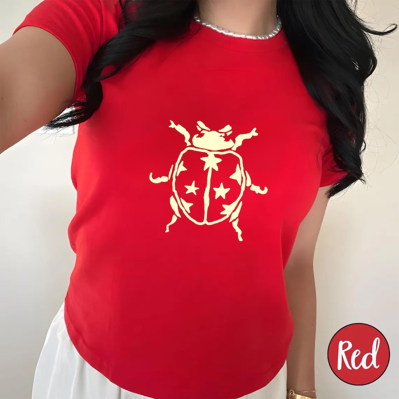 Op de afbeelding: Een felrood T-shirt met een cr&egrave;mekleurige lieveheersbeestjesafbeelding. Het lieveheersbeestjesontwerp heeft sterren op de rug. Het woord "Red" staat in een rode cirkel rechtsonder. Het shirt heeft korte mouwen.