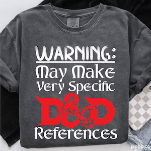 Könnte beinhalten: Dunkelgraues T-Shirt mit Rundhalsausschnitt und weißem Text: "WARNING: May Make Very Specific D&D References." Die D&D-Buchstaben sind rot mit einem Drachen-Design. Ein bequemer, lässiger Shirt-Stil.