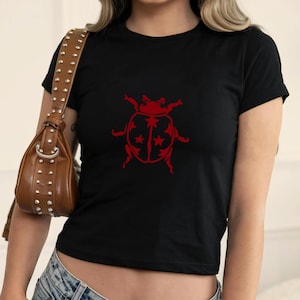 Op de afbeelding: Zwart T-shirt met een rode lieveheersbeestje-afbeelding. Het lieveheersbeestje-ontwerp heeft een gedetailleerde omtrek met steraccenten op de vleugels. Het shirt heeft korte mouwen en een ronde hals.