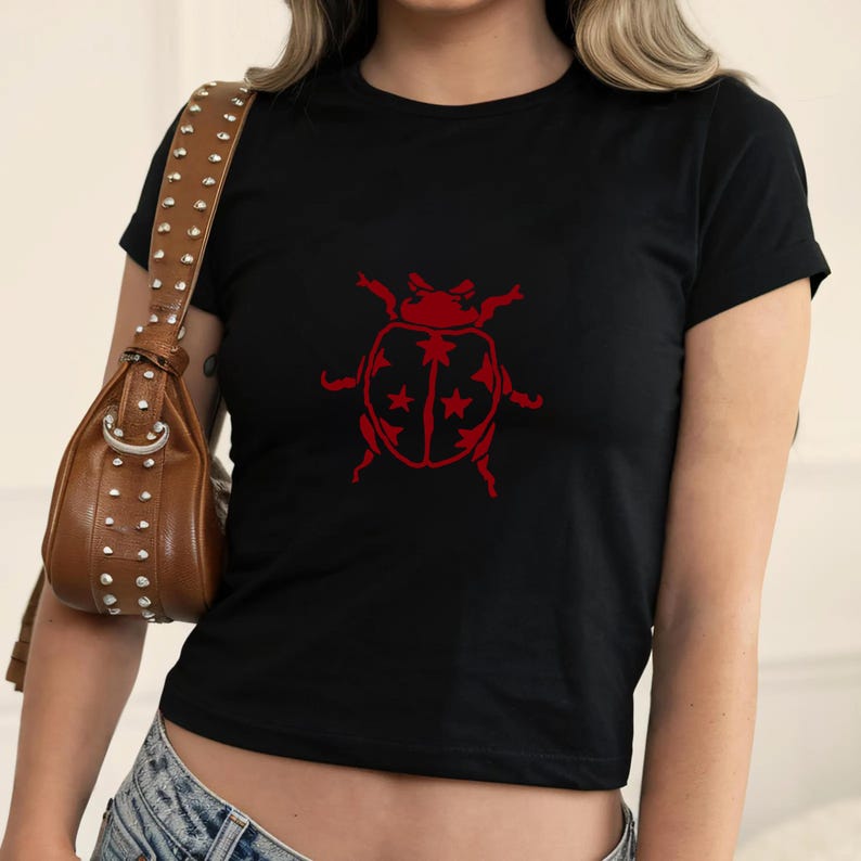 Op de afbeelding: Zwart T-shirt met een rode lieveheersbeestje-afbeelding. Het lieveheersbeestje-ontwerp heeft een gedetailleerde omtrek met steraccenten op de vleugels. Het shirt heeft korte mouwen en een ronde hals.