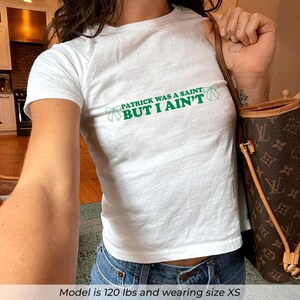 May include: White t-shirt with the text "PATRICK WAS A SAINT BUT I AIN'T" in green. The shirt has a crew neck and short sleeves. The text is in a playful font with a bow graphic.