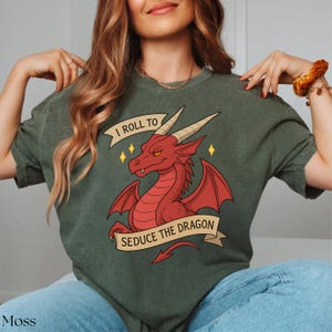 May include: Moss green t-shirt featuring a red dragon illustration with the text "I ROLL TO SEDUCE THE DRAGON". The dragon has horns, wings, and a tail. The shirt is a casual style.