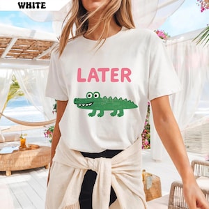 Pu&ograve; includere: T-shirt bianca con la scritta "LATER" in rosa sopra un alligatore verde a cartoni animati. L'alligatore ha i denti bianchi e il corpo verde. La maglietta &egrave; a girocollo.