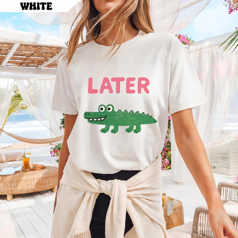 Pu&ograve; includere: T-shirt bianca con la scritta "LATER" in rosa sopra un alligatore verde a cartoni animati. L'alligatore ha i denti bianchi e il corpo verde. La maglietta &egrave; a girocollo.