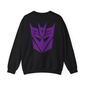 Könnte beinhalten: Schwarzes Sweatshirt mit einem großen lila Decepticon-Logo auf dem Rücken. Das Logo ist ein geometrisches Design mit scharfen Winkeln und einer zentralen augenartigen Form. Das Sweatshirt hat einen klassischen Rundhalsausschnitt.