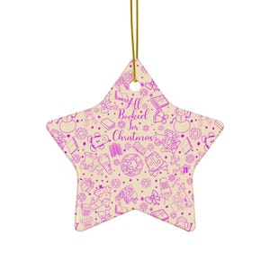 May include: Star-shaped Christmas ornament with a cream background and a repeating purple pattern of books, snowmen, and gifts. The text "All Booked for Christmas" is in the centre. A gold string is attached for hanging.