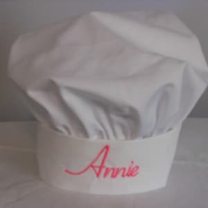 May include: A white chef's hat with a wide brim. The name "Annie" is embroidered in pink cursive on the brim. The hat is made of a soft, possibly cotton, material and is designed to be worn in a kitchen setting.
