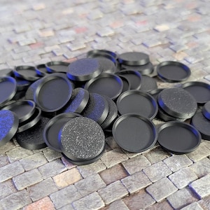 May include: A collection of numerous small, round, black plastic bases for miniature figures. Each base features a raised circular rim and a textured, slightly speckled surface, scattered in a pile on a gray stone-like surface.