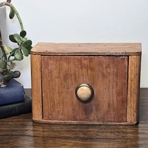 May include: A small, rectangular wooden drawer with a round, light-colored knob. The drawer is made of light brown wood with visible grain and is set on a dark wooden surface. A plant and books are in the background.