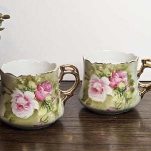May include: Two vintage teacups with gold handles and rims. The cups feature a hand-painted design of pink roses and green foliage on a light green background. The cups are displayed on a wooden surface.
