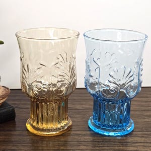 May include: Two vintage-style drinking glasses. One is amber-colored, the other is blue. Both glasses have a textured, embossed design and a pedestal base. The glasses are on a dark wooden surface.