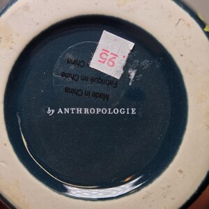 May include: Bottom view of a ceramic vase with a dark teal interior and a cream-colored rim. The base features the text "by ANTHROPOLOGIE" and a sticker with the number "25".