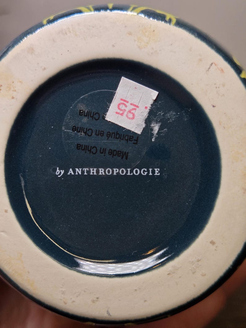 May include: Bottom view of a ceramic vase with a dark teal interior and a cream-colored rim. The base features the text "by ANTHROPOLOGIE" and a sticker with the number "25".