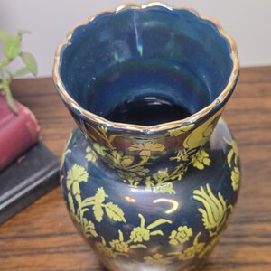 May include: A dark blue vase with a scalloped rim and gold trim. The vase features a floral pattern in a lighter shade of yellow. The vase is a decorative item.
