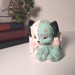 Puede incluir: Figura de perro de cerámica con un esquema de color verde menta y blanco. El perro tiene orejas negras, detalles florales rojos y amarillos y un lazo rosa. La figura está en una caja blanca.