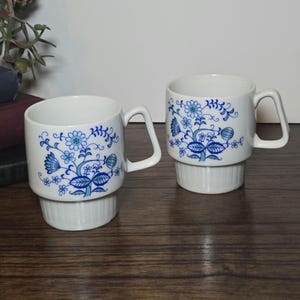 May include: Two white ceramic mugs with blue floral designs. The mugs have a cylindrical shape with a handle and sit on a ridged base. The blue floral pattern includes flowers and leaves. The mugs are on a wooden surface.