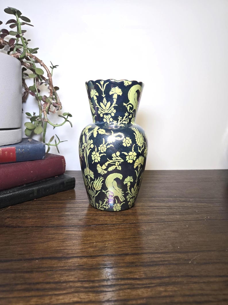 May include: A dark blue vase with a scalloped rim and a flared opening. The vase is decorated with a repeating pattern of yellow floral and bird motifs. The vase is sitting on a wooden surface.