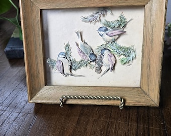 May include: A framed artwork featuring a detailed illustration of several small birds perched on evergreen branches. The birds have blue, purple, and white feathers. The frame is made of light brown wood and has a metal loop for hanging.