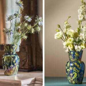 May include: A decorative, two-tiered vase with a dark blue and green iridescent finish. The vase is adorned with a floral pattern in yellow and holds a bouquet of flowers. The vase is displayed on a white surface.