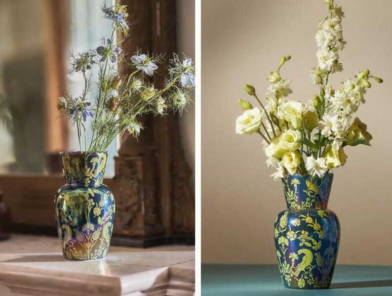 May include: A decorative, two-tiered vase with a dark blue and green iridescent finish. The vase is adorned with a floral pattern in yellow and holds a bouquet of flowers. The vase is displayed on a white surface.