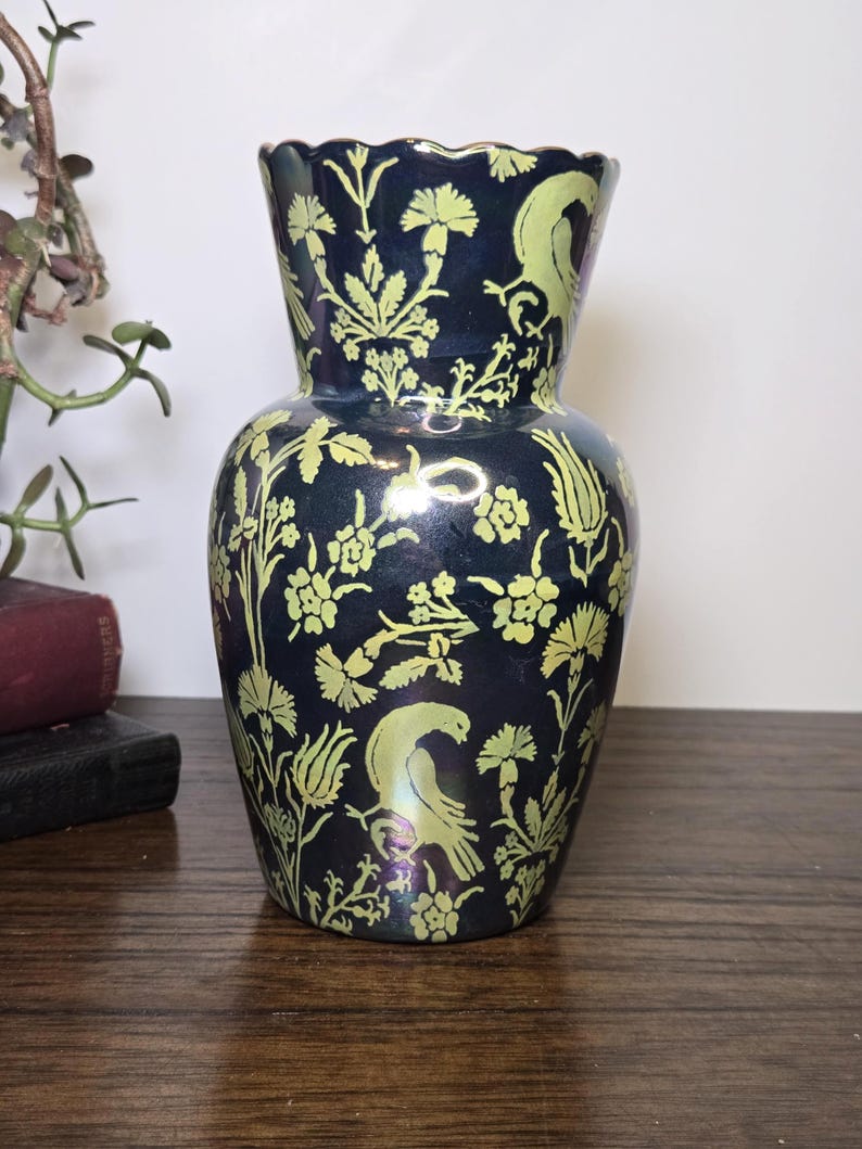 May include: A dark blue vase with a flared rim and a bulbous body. The vase is decorated with a repeating pattern of yellow-green floral and bird motifs. The vase is sitting on a wooden surface.