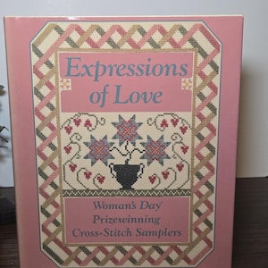 May include: A pink book titled "Expressions of Love" with cross-stitch samplers. The cover features a cross-stitch design with a floral motif and the text "Woman's Day Prizewinning Cross-Stitch Samplers."