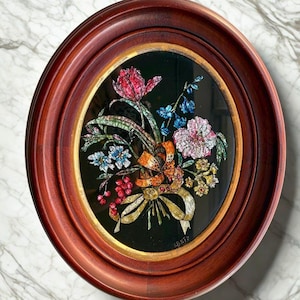 Peut inclure: Cadre ovale avec une œuvre florale. L'œuvre présente un bouquet de fleurs colorées, dont des fleurs rouges, bleues et roses, avec un ruban doré et un fond sombre. Le cadre est de couleur brun rougeâtre foncé.