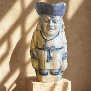Może przedstawiać: Ceramiczna figurka mężczyzny w niebieskim kapeluszu i szacie. Figurka jest biało-niebieska, z surowym wyrazem twarzy. Stoi na małym, kwadratowym, beżowym cokole. Tło jest w neutralnym kolorze z cieniami.