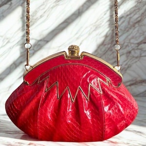 Può includere: Una borsa rossa a forma di conchiglia con una tracolla a catena dorata. La borsa ha una superficie strutturata e una chiusura dorata. Lo sfondo è una superficie simile al marmo con ombre.