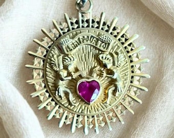 May include: Gold-toned pendant necklace with a sunburst design. The pendant features two figures holding a banner with text, a heart-shaped ruby, and intricate detailing. The necklace hangs on a silver chain.