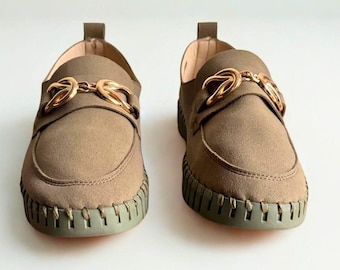 May include: A pair of olive green loafers with gold-coloured metal accents. The shoes have a rounded toe and a thick, textured sole. The loafers are displayed on a white background.