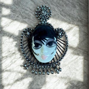 May include: A vintage silver-toned pendant featuring a painted face with black hair, large eyes, and pink lips. The face is set within an ornate, openwork frame with dangling bead accents. The pendant has a decorative top element.