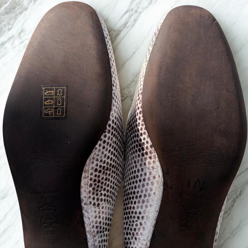 May include: A pair of women's flats with a snakeskin pattern in shades of white and gray. The soles are dark brown with the brand name and size information visible. The shoes are displayed on a marble surface.