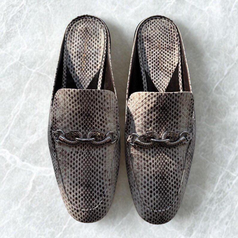 May include: A pair of gray and brown snakeskin-patterned slip-on loafers. Each shoe features a silver chain detail across the vamp. The shoes have a pointed toe and a low heel.