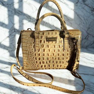 Puede incluir: Bolso de mano color beige con un patrón en relieve similar al de un cocodrilo. El bolso tiene un asa superior y una correa para el hombro desmontable. Una pequeña placa dorada con el nombre de la marca está centrada en la parte delantera. El bolso está sobre una superficie de mármol.