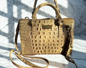 May include: Tan handbag with a crocodile-style embossed pattern. The bag features a top handle and a detachable shoulder strap. A small gold-coloured plaque with the brand name is centered on the front. The bag is on a marble surface.