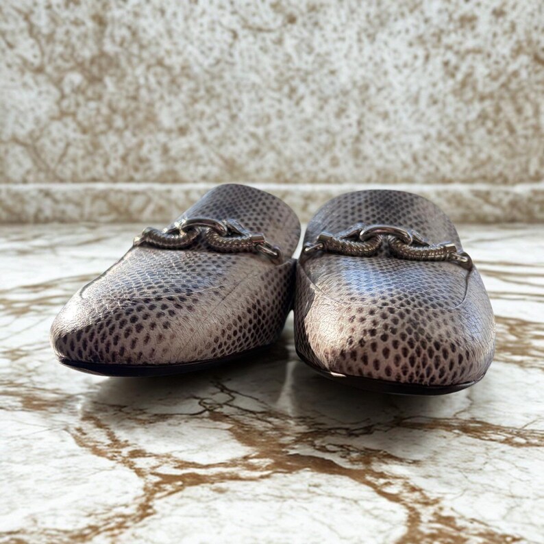 May include: A pair of slip-on shoes with a snakeskin pattern in shades of gray and brown. Each shoe has a decorative silver-toned chain across the top. The shoes are displayed on a marble surface.
