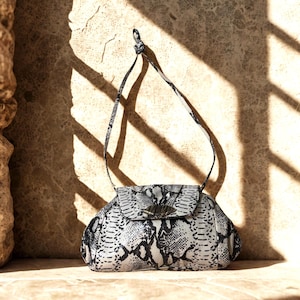 May include: A small, black and white snakeskin-patterned handbag with a flap closure and a shoulder strap. The bag has a silver-toned clasp and is set against a textured, neutral-toned background.
