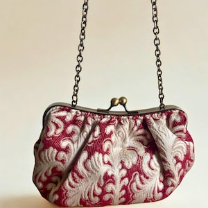 May include: A red and silver patterned clutch with a chain strap. The clasp is a dark bronze color. The bag has a decorative floral design.