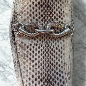May include: Close-up of a snakeskin-patterned shoe. The shoe features a silver metal accent with a horse head design. The snakeskin pattern is a mix of brown, gray, and cream colors.