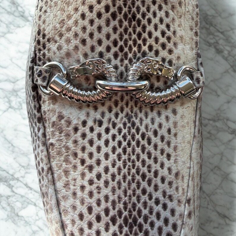 May include: Close-up of a snakeskin-patterned shoe. The shoe features a silver metal accent with a horse head design. The snakeskin pattern is a mix of brown, gray, and cream colors.