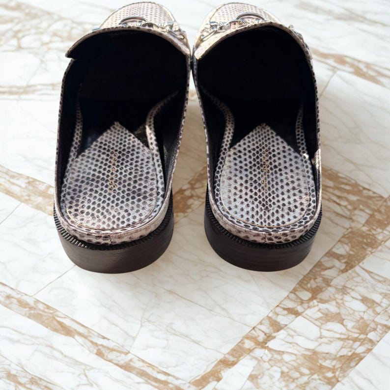 May include: A pair of backless loafers with a snakeskin pattern in shades of beige and black. The shoes have a black sole and a silver chain detail across the top. The interior of the shoe is also patterned.
